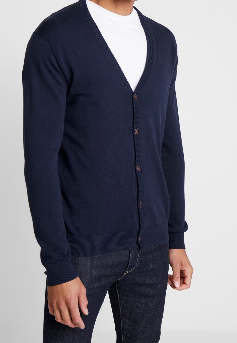 Homme portant un cardigan bleu marine boutonné sur un t-shirt blanc et un jean bleu foncé sur un fond uni.
