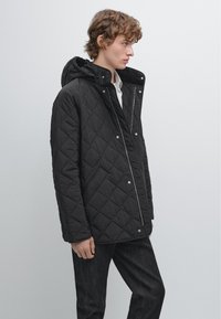 Sort quilted jakke med hætte, der har trykknapper og diagonale syninger. Modellen har den på over en hvid skjorte og mørke jeans.