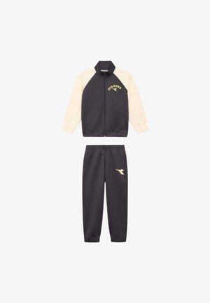Completo sportivo nero e crema caratterizzato da una giacca con zip e un logo Diadora dorato, accompagnato da pantaloni coordinati con un accento del logo sulla gamba.