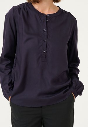 Woman wearing a long-sleeve navy blouse with a round collar and four-button placket, hands in pockets, paired with black pants.