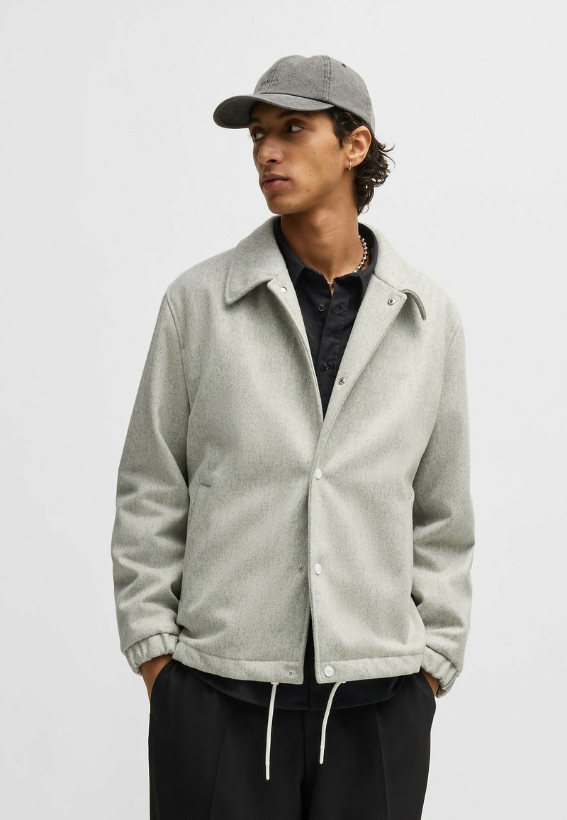 Light grey wool blend jacket with a collared neckline, snap buttons, and elastic cuffs. Paired with a dark shirt and grey cap.