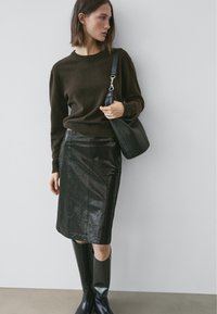 Pull marron, jupe-crayon en cuir noir brillant, bottes noires montantes jusqu'aux genoux, et sac à main noir texturé, le tout sur un fond gris clair.