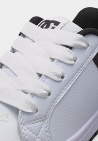 Primo piano di una sneaker bianca con lacci bianchi spessi, fodera in tessuto nera e logo nero sulla linguetta imbottita.