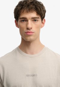 Beige bomulds t-shirt med korte ærmer og rund halsudskæring, med diskret "SUPERDRY" logo broderet i matchende tråd på brystet.