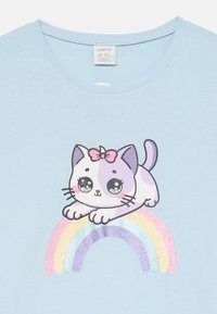 Hellblaue Baumwoll-T-Shirt mit einem grafischen Druck einer lila und weißen Katze mit einer rosa Schleife, die auf einem glitzernden Regenbogen sitzt.