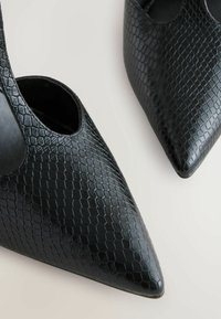 Svarta högklackade skor med spetsig tå, tillverkade av textilmaterial med ett bikake-mönster, öppna sidor och en elegant design.