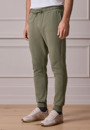Uomo che indossa pantaloni jogger verde oliva con coulisse in vita, scarpe da ginnastica bianche e calzini scuri, in piedi su un pavimento in legno chiaro all'interno.
