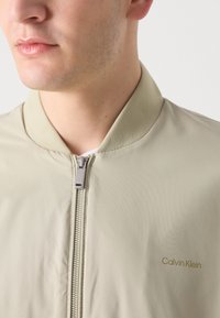 Hellbeige Bomberjacke mit ribbeltem Kragen, Frontreißverschluss und goldfarbenem Logo-Akzent. Glatte Textur und minimalistisches Design.