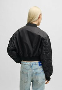 Svart bomberjacka med ribbad mudd och krage, med en slät, vadderad textur. Bärs med ljusblå jeans.