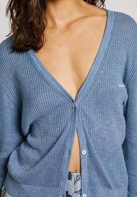 Cardigan rigato di un blu chiaro con scollatura a V profonda, chiusura con bottoni e maniche lunghe; presenta una texture morbida e un delicato dettaglio del logo.