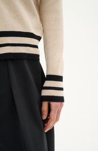 Pull à côtes beige avec des accents de rayures noires, présentant des poignets larges. Le tissu semble doux avec une texture subtile. Jupe noire partiellement visible.