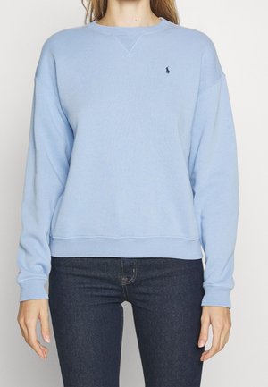Femme portant un sweat-shirt bleu clair à manches longues avec un petit logo sombre sur la poitrine et un jean bleu foncé, debout devant un fond uni.