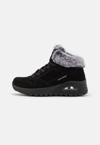 Zapatillas deportivas de ante negro con un collar de piel sintética, suela gruesa y característica de bolsillo de aire. Diseño con cordones y marca "Skechers" en el lateral.