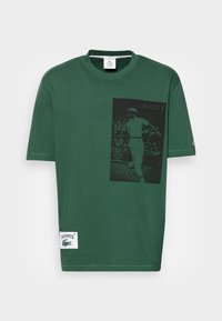 Camiseta de algodón verde con un gráfico negro de un jugador de tenis y el logo "LACOSTE". Incluye una etiqueta de Lacoste en el dobladillo.