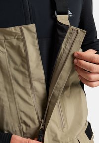 Bežna izolirana delovna obleka z zadrgami in žepi, s črnim notranjim delom, nastavljivimi trakovi in gladko, trpežno teksturo tkanine.