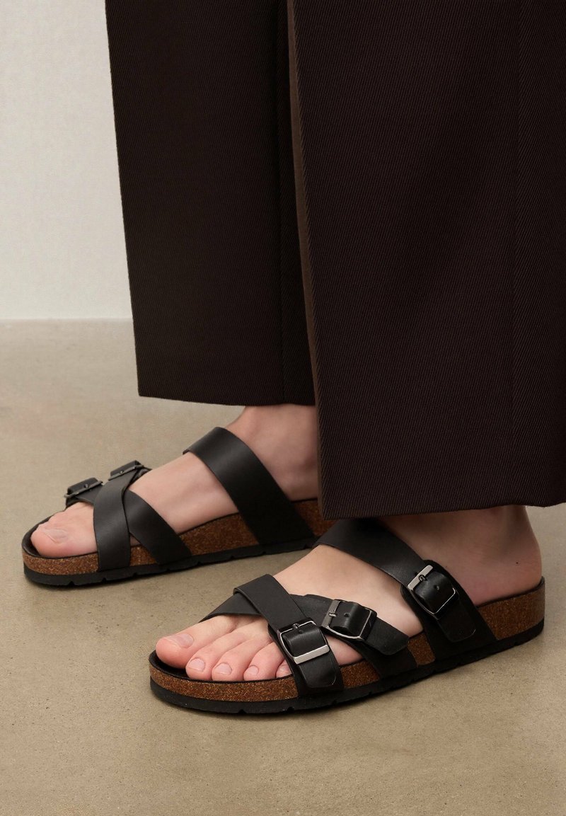 Pieds portant des sandales en cuir noir avec trois brides réglables à boucle et des semelles en liège, assorties à un pantalon large et sombre.