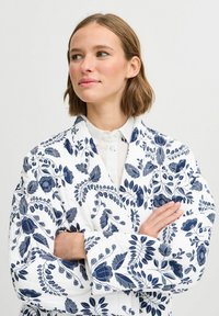 Balts dīda jaku ar tumši zilu ziedu rakstu, kurai ir šaura apkakle un garas piedurknes, izstrādāta ar sarežģītiem lapu un ziedu motīviem.