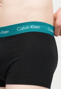 Zbliżenie na osobę noszącą czarne bokserki Calvin Klein z turkusowym pasem z napisem "Calvin Klein".