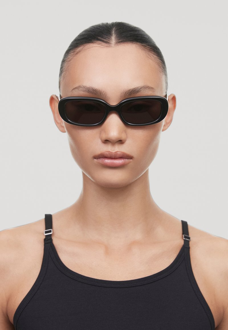 Ochelari de soare ovali negri cu lentile întunecate, având o ramă groasă din plastic. Modelul poartă un top negru cu bretele ajustabile.