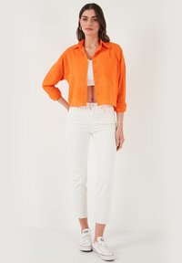 Orange linen cropped shirt with button closure and pocket, paired with white skinny jeans and white sneakers. Smooth, lightweight fabric.