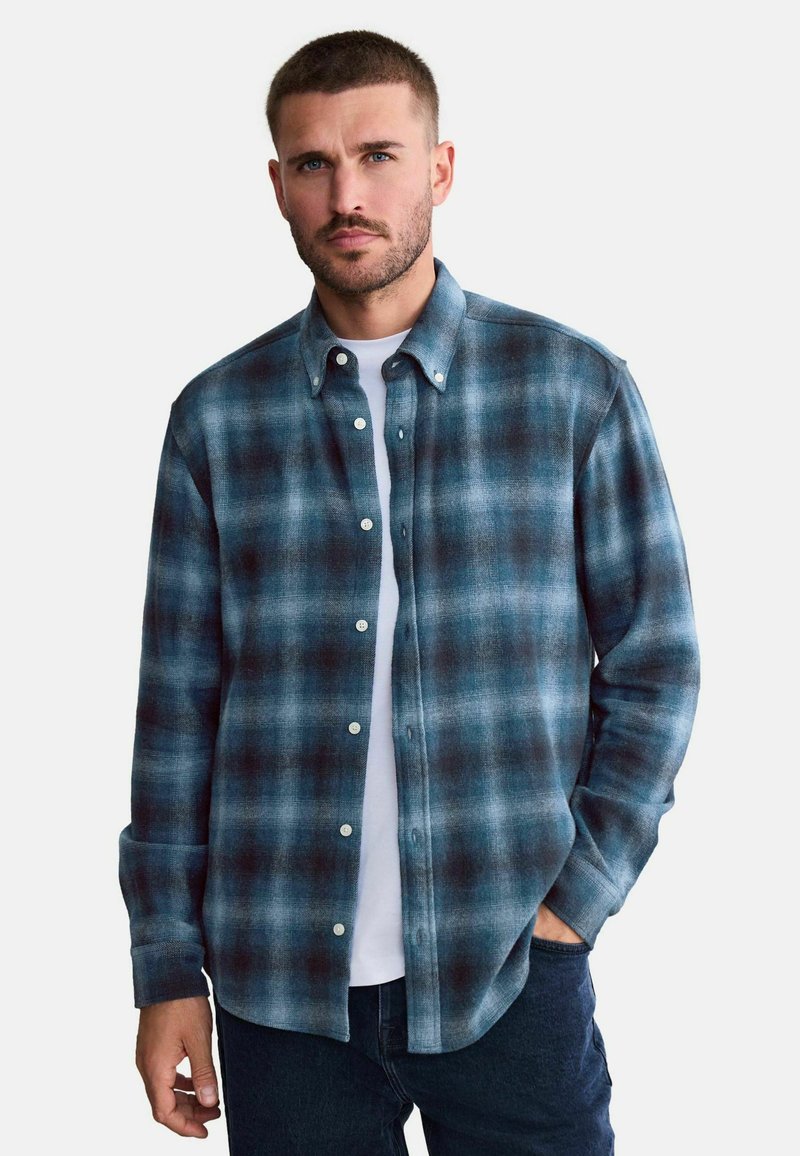 Homme portant une chemise à carreaux bleue et noire boutonnée sur un t-shirt blanc avec un jean foncé, debout avec une main dans la poche, expression neutre.