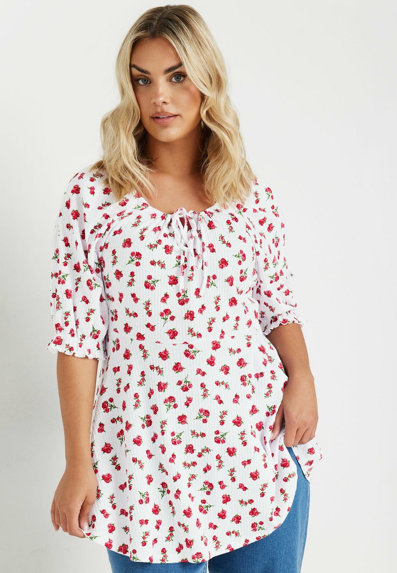Blusa blanca con pequeño estampado de flores rojas, cuello fruncido con cordón y mangas abullonadas de tres cuartos. Tejido ligero y texturizado.