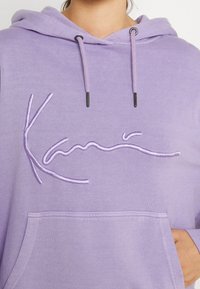 Lila hoodie med dragsko, med broderad text i en ljusare lila nyans. Inkluderar en framficka och mjuk textur.