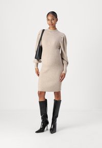 Beige ribbestrikket kjole med lange puffermer, knelength, kombinert med svarte knelange stiletto støvler og en liten svart veske.