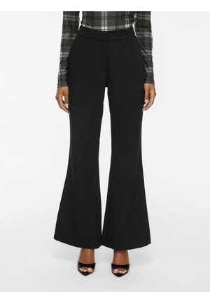 Femme portant un pantalon noir évasé, des talons ouverts noirs et un haut gris à carreaux à manches longues, debout devant un fond blanc.