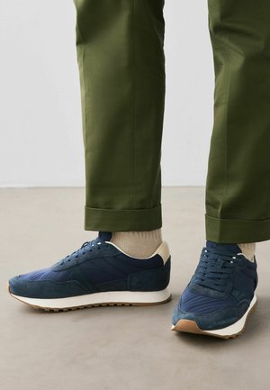 Feet wearing navy blue and beige sneakers with white soles, paired with beige socks and olive green cuffed trousers on a neutral floor.