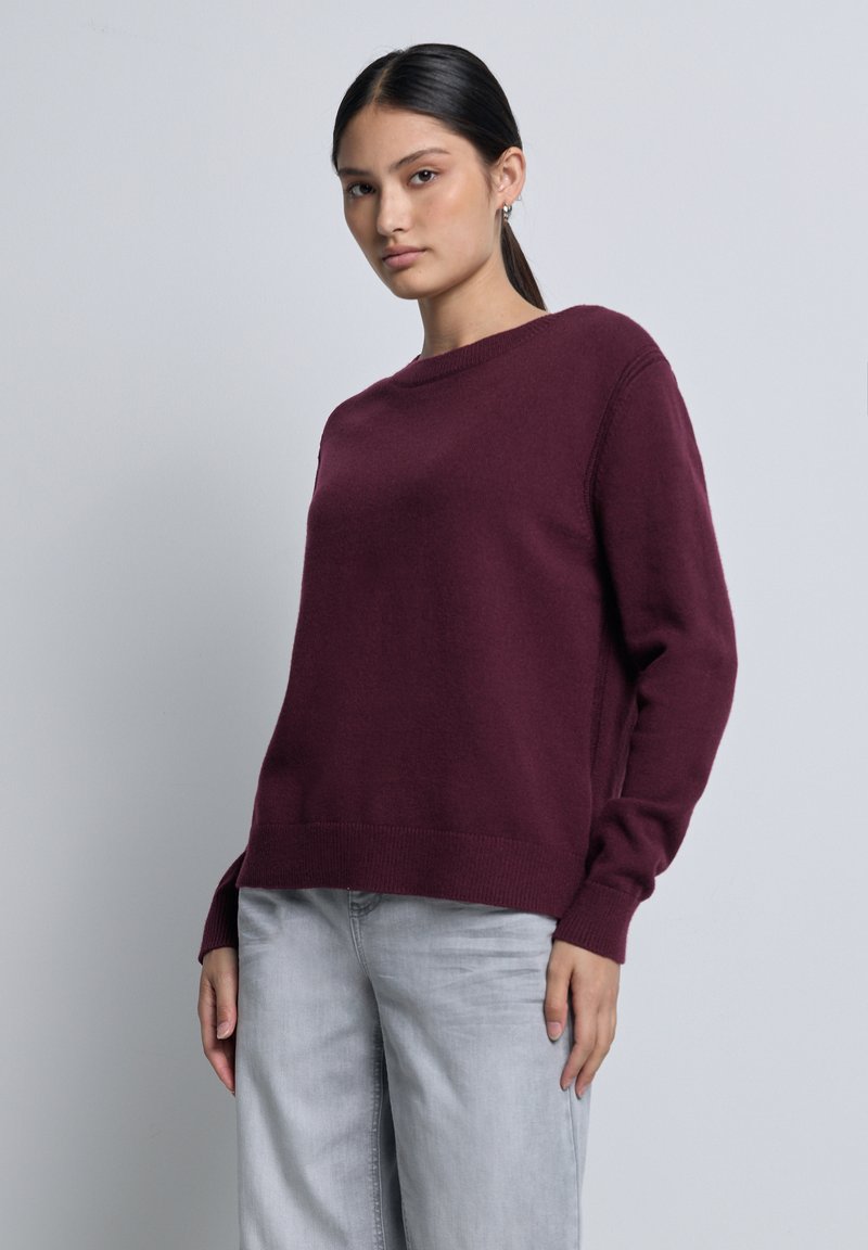 Femme aux cheveux bruns en queue de cheval portant un pull à manches longues bordeaux et un jean gris clair sur un fond clair et uni.