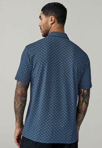 Camisa de manga corta en tela azul oscuro con un patrón texturizado que recuerda a formas de abanico superpuestas. Incluye un collar clásico y un corte relajado.