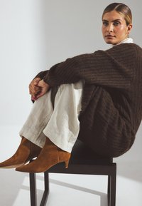 Pull en maille marron, pantalon crème et bottines à talon aiguille en suedine marron. Modèle assis sur une chaise noire devant un fond clair.