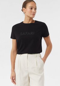 Czarna bawełniana koszulka z napisem "SAFARI" w błyszczącym wykończeniu. Okrągły dekolt, krótkie rękawy, luźny krój, zestawiona z jasnobeżowymi spodniami.