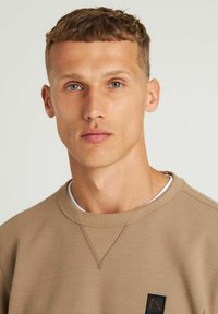 Sudadera beige con cuello redondo de canalé y costuras reforzadas, que presenta un pequeño parche de logo negro en el frente. Textura suave.