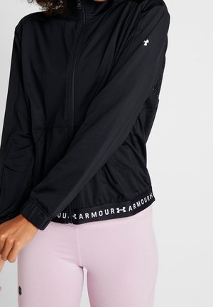 Personne portant une veste zippée noire Under Armour et des leggings rose clair sur un fond clair uni.