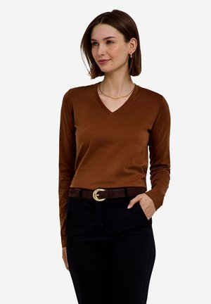 Femme aux cheveux courts et bruns, portant une chemise marron à manches longues et col en V, et un pantalon noir avec une ceinture foncée, debout avec une main dans la poche.