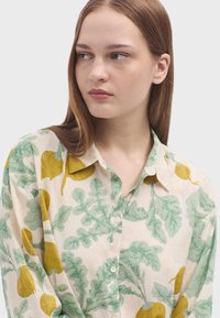 Camicia leggera di colore crema con motivi di foglie verdi e radici gialle, dotata di bottoni sul davanti e colletto.