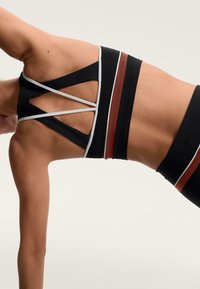 Čierna športová podprsenka so širokými ramienkami, s bielymi a hrdzavočervenými akcentovými pruhmi. Sieťovinové detaily a krížový dizajn na chrbte.