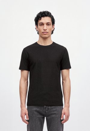 Un homme se tient face à l’objectif, vêtu d’un T-shirt noir à manches courtes et à la texture marquée, ainsi que d’un jean gris foncé, sur un fond clair et uni.