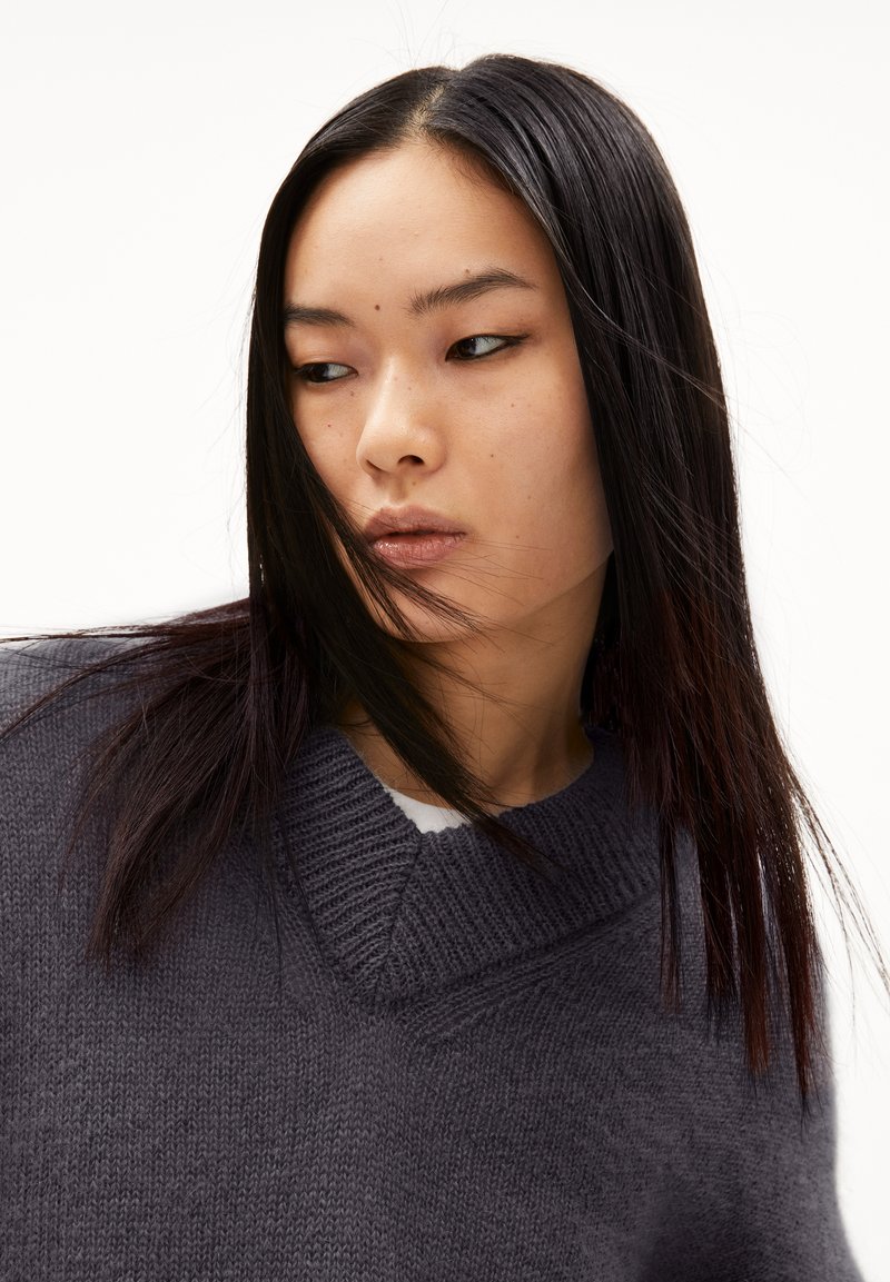 Chunky donkergrijze gebreide trui met een V-halsontwerp en ribbelachtige halslijn. Glad materiaal met zichtbaar breipatroon. Haar bedekt gedeeltelijk het gezicht.