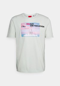 Camiseta de algodón verde claro con un gráfico rectangular que presenta un coche, un edificio y el texto "HUGO ON VACATION" en negro y rojo.
