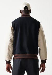 Ανδρικό μπουφάν με σώμα από μάλλινο ύφασμα σε ναυτικό μπλε χρώμα, μανίκια από κρεμ δέρμα και καφέ ριμπ μανσέτες. Διαθέτει στρογγυλεμένο γιακά και ελαφρώς κοντό μήκος.