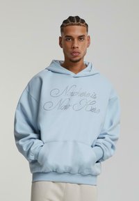 Gaiši zils hoodie ar izšūtu tekstu "Tagad nekur nav šeit", ribotām piedurknēm un priekšējo kangaroo kabatu. Mīksts audums ar brīvu piegriezumu.