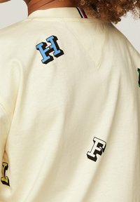 Cremefarvet sweatshirt med flerfarvede bogstaver (H, F, L) i forskellige skrifttyper spredt over ryggen. Bomuldsstof med en glat tekstur.