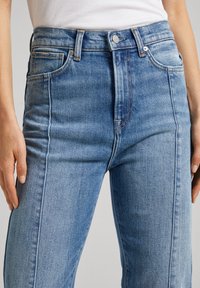 Jeans de denim azul de cintura alta con una textura suave, que presentan costuras naranjas y un cierre frontal con botón, diseñados para un ajuste ceñido.