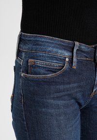 Mörkblå denimjeans med slim fit, med orange sömnad, en liten framficka och en slät textur. Kanterna är lätt slitna.