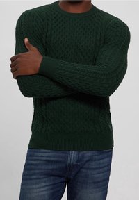 Dark green cable-knit sweater with a round neckline, ribbed cuffs, and hem. Worn over blue jeans, featuring a textured fabric pattern.