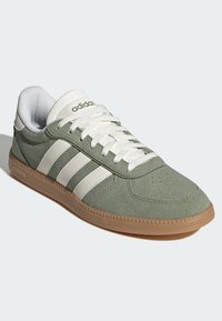 Zeleni sneaker od brušene kože s bijelim kožnim prugama, gumeni potplat i vezice. Opremljen je mekanom ovratnikom i teksturiranom kapicom na vrhu.