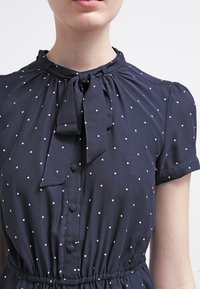 Mörkblå klänning med vita prickar, korta ärmar, en slipskrage och knäppning fram. Rynkad midja för en figurnära siluett.